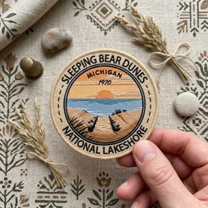 May include: A circular embroidered patch featuring a landscape of Sleeping Bear Dunes in Michigan, dated 1970. The design depicts a sunset over a blue lake with sandy dunes in the foreground, all enclosed by a black border with text.
