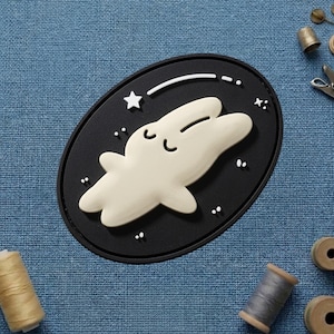 May include: An oval-shaped, black patch featuring a cream-colored cartoon dolphin with a smiling face. A white star and a curved line with dots are above the dolphin. The patch is on a blue textured surface.