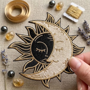 May include: An embroidered patch featuring a sun and moon design with a black sun face and a cream-colored crescent moon face. The design includes gold-colored thread detailing and sharp, pointed rays extending outward from the central celestial figures.