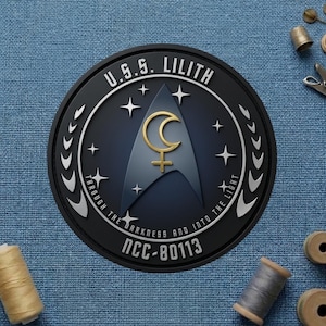 May include: A circular black patch with the text "U.S.S. LILITH" and "THROUGH THE DARKNESS AND INTO THE LIGHT". The patch features a gold crescent moon and a symbol on a blue background, surrounded by stars. The text "NCC-80113" is also present.