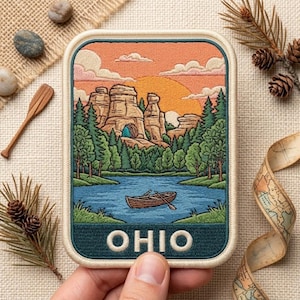May include: A rectangular embroidered patch featuring a landscape scene with a blue lake, a wooden rowboat, green pine trees, and tall rock formations under a sunset sky. The word OHIO is stitched in white capital letters at the bottom.