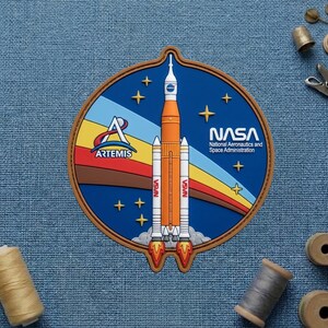 May include: A circular patch featuring the NASA logo and the Artemis program emblem. The design includes a rocket launching, stars, and a rainbow-colored arc. The patch has a brown border and the text "NASA National Aeronautics and Space Administration".