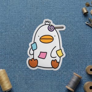 May include: A white duck-shaped patch with a yellow beak and orange feet. The duck features a purple and white spiral lollipop graphic on its head and three small rectangular sticky notes in blue, pink, and yellow attached to its body.