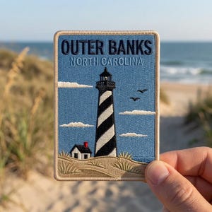 May include: A rectangular embroidered patch featuring a black and white spiral-striped lighthouse, a small house with a red chimney, and sand dunes. The text reads OUTER BANKS NORTH CAROLINA in dark blue against a light blue sky with white clouds.