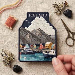May include: A rectangular embroidered patch with a dark blue border featuring a landscape of Iceland. The design includes a mountain range, white clouds, a harbor with colorful houses, and two boats on blue water. The word ICELAND is stitched at the top.