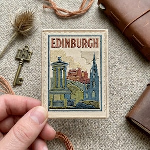 May include: A rectangular embroidered patch featuring the word EDINBURGH in brown text at the top. The design depicts a blue monument, a red building, and a blue church spire against a cream background, all enclosed within a stitched border.