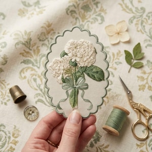 May include: An oval-shaped fabric patch with a scalloped edge featuring an embroidered design of white hydrangea flowers with green leaves and a tied ribbon. The embroidery is centered on a cream-colored background with a sage green border.