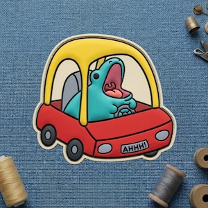 May include: A whimsical, cartoon-style patch featuring a teal hippo with an open mouth, inside a red car with a yellow roof. The car has black wheels and the text "AHHH!" on the front.