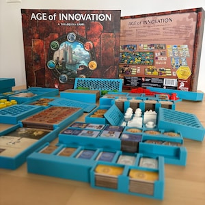 Può includere: Gioco da tavolo "Age of Innovation" con una scatola che presenta un design circolare e testo. I componenti del gioco, tra cui carte e gettoni, sono organizzati in vassoi blu. Il gioco è ambientato su una superficie di legno.