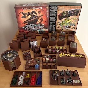 Può includere: Un set di gioco da tavolo Mage Knight Ultimate Edition con numerosi organizzatori, portacarte e vassoi in plastica marrone. Il set include vari componenti di gioco, miniature, cristalli colorati e gettoni circolari disposti in ordine.