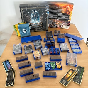 Può includere: Un set di giochi da tavolo, "Syncanite Foundation", con vari componenti disposti su una superficie di legno. Il set include una scatola, carte, vassoi blu, pedine e sacchetti. La scatola presenta il titolo e l'artwork del gioco.
