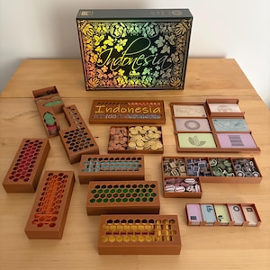 Op de afbeelding: Het bordspel "Indonesia" met een holografische doos en diverse houten spelonderdelen. De spelcomponenten omvatten houten trays, gekleurde tokens, kaarten en een doos met de speltitel.