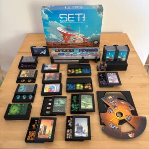 May include: A board game titled "SETI: Search for Extraterrestrial Intelligence" with a box featuring a blue sky and radio telescopes. The game includes various components like cards, tokens, and a circular game board, all arranged on a wooden surface.