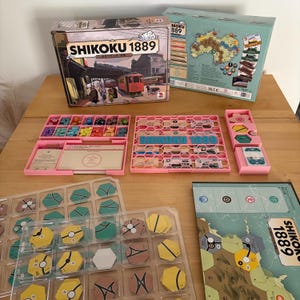 Può includere: Gioco da tavolo "Shikoku 1889" con una scatola che presenta una scena di treno d'epoca. Il gioco include vassoi rosa, pedine e una mappa. Le pedine sono di vari colori.