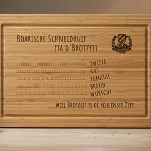 Könnte beinhalten: Rechteckiges Schneidebrett aus Bambus mit graviertem Text und dekorativem Emblem. Der Text enthält deutsche Sätze. Das Brett hat eine vertiefte Kante und ist für die Zubereitung von Speisen konzipiert.