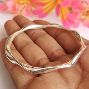 Może przedstawiać: Polerowana srebrna bransoletka typu bangle o ciągłym, falistym wzorze. Metalowa powierzchnia jest gładka i odblaskowa, chwytając światło wzdłuż zakrzywionych, nieregularnych krawędzi, tworząc płynny, organiczny kształt.