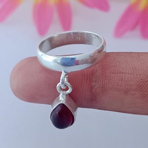Op de afbeelding: Een zilveren ring met een druppelvormige, dieprode edelsteen hanger. De ring wordt door een vinger vastgehouden, met een vage achtergrond van roze en gele bloemen.