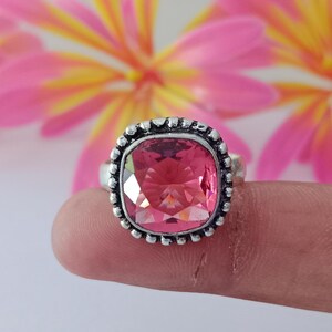 Könnte beinhalten: Ein silberfarbener Ring mit einem großen, quadratisch geschliffenen, facettierten rosa Edelstein, der in einen dekorativen, perlenartigen Metallrand eingefasst ist. Der Ring hat ein poliertes, strukturiertes Finish um die Fassung.