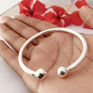 May include: A polished silver-tone cuff bracelet featuring a smooth, open-ended design with a spherical ball bead at each terminal end. The metal surface has a reflective, metallic finish that catches the light, creating a clean and simple aesthetic.