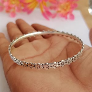 May include: A silver bangle bracelet with a floral design. The bracelet is round and features a repeating pattern of small, detailed flowers around the band. The metal has a polished, reflective surface, and the bracelet is shown on a hand.