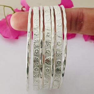 May include: A set of six silver bangles, each with a textured surface and engraved geometric patterns including starbursts and star shapes. The bangles are held against a backdrop of pink flowers.