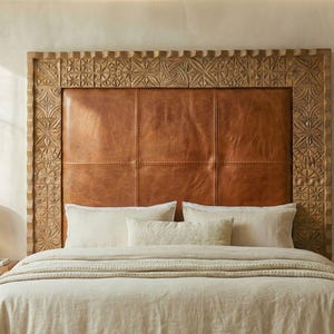 May include: A bed featuring a large, carved wooden headboard with a brown leather panel. The bed has white bedding, pillows, and a beige blanket. A lamp sits on a wooden side table. The room has a neutral colour palette.
