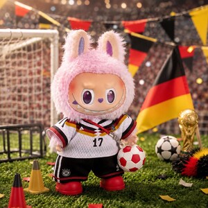 May include: A plush toy character with pink fur and rabbit ears wearing a white and black soccer jersey with the number 17, black shorts, and red shoes. The character holds a small soccer ball and stands on a green field with soccer gear.