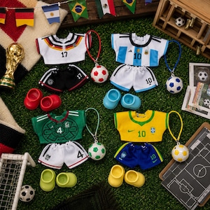 May include: Four sets of doll-sized soccer uniforms including jerseys, shorts, and shoes, featuring the national team colors and logos of Germany, Argentina, Mexico, and Brazil, accompanied by four miniature soccer ball keychains.