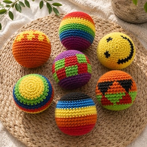 Può includere: Una collezione di sette palline all'uncinetto colorate. Le palline presentano vari disegni, tra cui un motivo arcobaleno, una faccina sorridente e forme geometriche. Le palline sono realizzate con filato nei toni del rosso, arancione, giallo, verde, blu e viola.