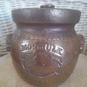 May include: Brown stoneware crock with a lid. The crock has two handles and the word "MONBULK" is embossed on the front. The crock has a decorative band around the top and a small knob on the lid.