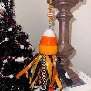 May include: A hanging ornament shaped like a candy corn with a white top, orange middle, and yellow bottom. It features a beaded string with orange and white stripes and yellow beads, finished with a cluster of black, orange, and yellow ribbons.