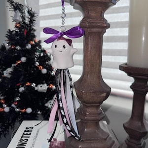May include: A white ceramic ghost ornament featuring a small purple bow on its head, suspended by a clear beaded string. A larger purple ribbon bow sits above the ghost, and a cluster of black, white, and pink ribbons hangs below it.