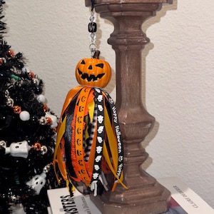 May include: A hanging Halloween ornament featuring a carved orange jack-o'-lantern head attached to a cluster of ribbons in black, orange, and yellow. The ribbons include patterns of ghosts and pumpkins, with one ribbon printed with the text Happy Halloween.