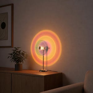 Puede incluir: Una lámpara de escritorio moderna, en tono dorado, proyecta un efecto de puesta de sol colorido en la pared. La lámpara presenta un diseño circular con una fuente de luz central y una serie de discos translúcidos concéntricos. Crea un ambiente cálido.