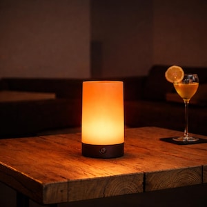 May include: A cylindrical, orange-glowing lamp with a dark base and a power button sits on a wooden table. A cocktail with a lemon slice garnish is in the background. The lamp emits a warm, inviting light, creating a cozy ambiance.