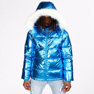 May include: A shiny, metallic blue puffer jacket with a hood trimmed with white faux fur. The jacket has a zipper closure and the text "VENGADE71" is visible on the chest. The model is wearing light blue jeans.