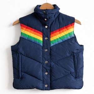 May include: A navy blue quilted vest with a rainbow stripe across the chest. The vest has a high collar, a button-up front, and two pockets. The vest is hanging on a wooden hanger.