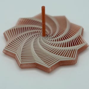 May include: A geometric, star-shaped object with a central, vertical peg. The design features layers of white and peach-colored lines, creating a spiral effect. The object is likely a decorative or functional item.