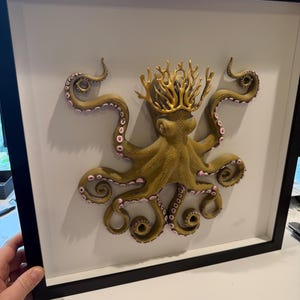 May include: A framed, three-dimensional artwork featuring an octopus with a gold crown. The octopus is olive green with pink accents on its tentacles. The artwork is set against a white background within a black frame.