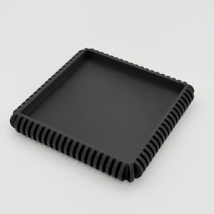 May include: A square, black tray with a ribbed edge. The tray has a slightly textured surface and a raised, decorative border. The design is simple and modern, suitable for various uses.