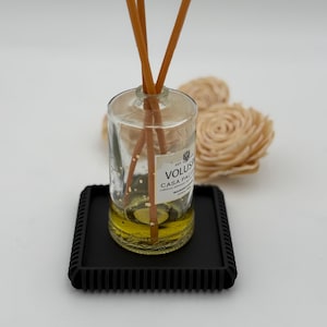 May include: A clear glass diffuser bottle with brown reeds, on a black square tray. The bottle contains a yellow liquid and has a label that reads "VOLUSPA CASA PACIFICA". Two decorative beige flower-shaped objects are in the background.