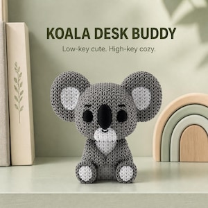 May include: A gray and white crocheted koala figurine with large round ears, black eyes, and a black nose. The text above reads KOALA DESK BUDDY and Low-key cute. High-key cozy. The koala has a white chest patch and white accents on its feet and ears.