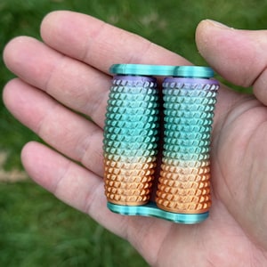 May include: A multi-colored, textured, cylindrical object with a connecting base. The colors transition from orange to teal to purple. The object is held in a person's hand against a blurred green background. The object appears to be 3D printed.