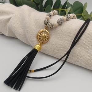 Peut inclure: Un collier avec un pendentif soleil doré et un pompon noir. Le collier a une chaîne de perles avec des perles crème et tachetées. Le pompon est en matière noire semblable à du daim.