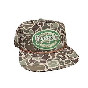 May include: A camouflage pattern baseball cap with a brown rope detail. The cap features a green oval patch with the text "Remington" and "America's Oldest Gunmaker". The hat is a classic accessory.