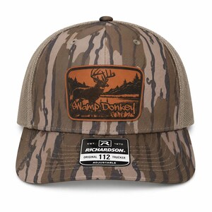 May include: A camouflage trucker hat with a brown leather patch featuring a buck deer illustration and the words "Swamp Donkey." The hat has a mesh back and a curved brim. The Richardson brand label is visible.