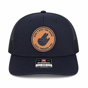 May include: Navy blue trucker hat with a black mesh back and a brown leather patch. The patch features a hand gesture and the text "DON'T BITE THE HAND THAT FINGERS YOU". The hat has an adjustable strap and a Richardson tag.