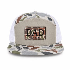 May include: A camouflage-patterned baseball cap with a white mesh back. The front panel is tan with a rectangular patch that reads "DAD EST. 2026" in black lettering. The brim features a matching camouflage design.
