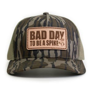 May include: A camouflage trucker hat with a brown leather patch. The patch features the text "BAD DAY TO BE A SPIKE" in brown lettering, along with a deer graphic. The hat has a mesh back and a curved brim.