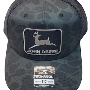 May include: A black and green camouflage trucker hat with a mesh back. The front features a rectangular patch with the John Deere logo and text. The hat is adjustable and has a Richardson 112 Trucker tag.